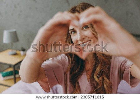Young woman wear purple t-shirt pajama lying in bed showing shape heart look through hands heart-shape sign rest spend time in bedroom lounge home in own room hotel wake up dream be in good mood day