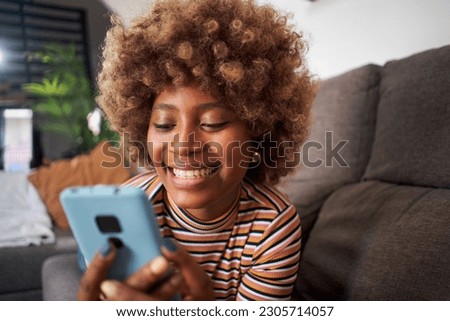 Close up of smiling African American pretty woman using cell phone lying on sofa in living room. Happy young black afro girl flirting on mobile dating app at home. People looking for life partner. Royalty-Free Stock Photo #2305714057
