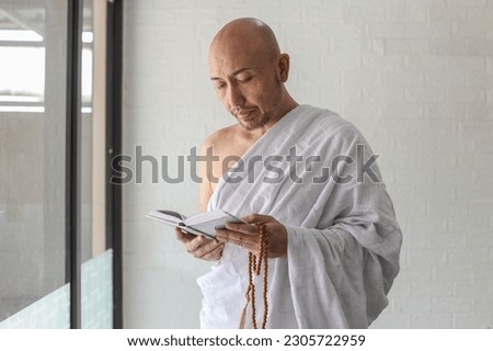Muslim man wearing traditional white ihram clothes, reading quran while holds prayer beads.  Royalty-Free Stock Photo #2305722959