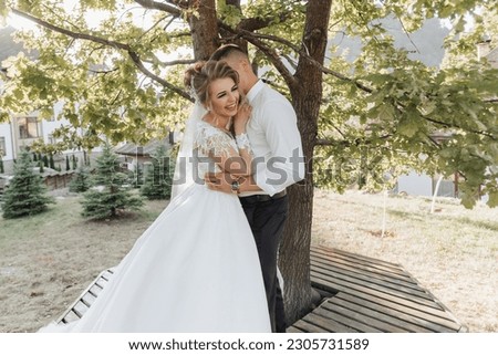 Wedding portrait. A groom in a black suit and a blonde bride are standing, hugging and kissing under a tree. Photo session in nature. A beautiful ray of the sun in the photo. Beautiful hair and makeup