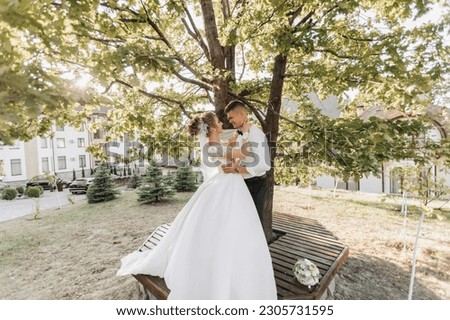 Wedding portrait. A groom in a black suit and a blonde bride are standing, hugging and kissing under a tree. Photo session in nature. A beautiful ray of the sun in the photo. Beautiful hair and makeup