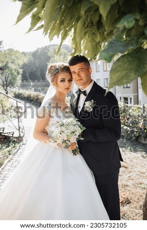 Wedding portrait. A groom in a black suit and a blonde bride stand embracing by a stone wall under a tree. Photo session in nature. Beautiful hair and makeup