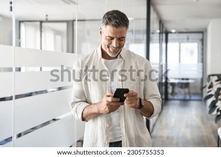 Smiling mature Latin or Indian businessman holding smartphone in office. Middle aged manager using cell phone mobile app. Digital technology application and solutions for business success development.