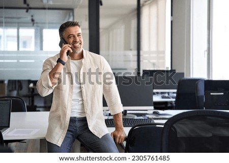 Middle aged Latin or Indian businessman having call on smartphone with business partners or clients. Smiling mature Hispanic man in relaxing pose talking by mobile cellphone at work in modern office.  Royalty-Free Stock Photo #2305761485