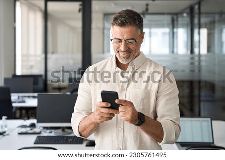 Close-up of smiling mature Latin or Indian businessman holding smartphone in office. Middle aged manager using cell phone mobile app. Digital technology application and solutions for business concept. Royalty-Free Stock Photo #2305761495