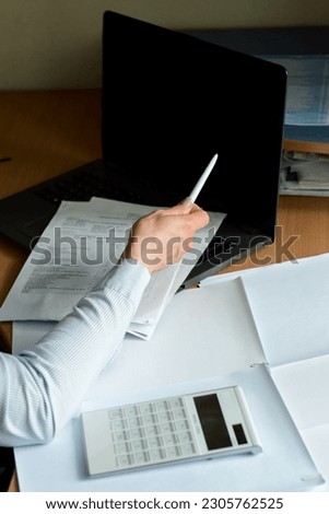 Woman's right hand with a pen in it is pointing at the blank black screen of laptop, blank documents and white calculator are lying near on the desktop. Front view