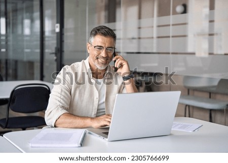 Middle aged Latin or Indian businessman having call on smartphone with business partners or clients. Smiling mature Hispanic man sitting at table talking by mobile cellphone at workplace in office.  Royalty-Free Stock Photo #2305766699