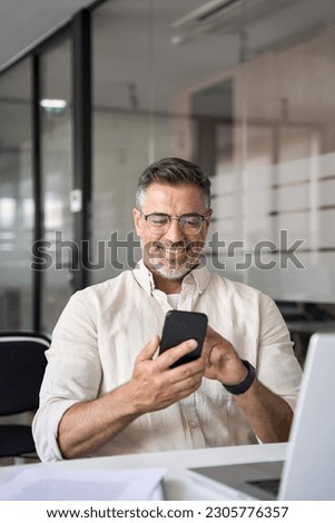 Vertical portrait of middle aged Hispanic business manager using cell phone mobile app. Smiling Latin or Indian mature man businessman holding smartphone sit in office working online on gadget.