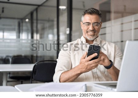 Middle aged Hispanic business manager ceo using cell phone mobile app, laptop. Smiling Latin or Indian mature man businessman holding smartphone sit in office working online on gadget with copy space. Royalty-Free Stock Photo #2305776359