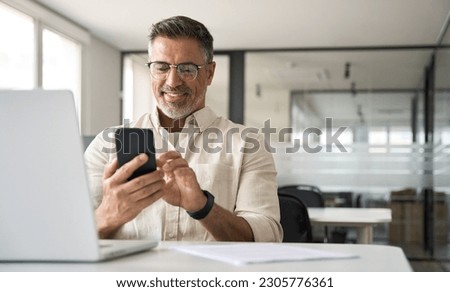 Middle aged Hispanic business manager ceo using cell phone mobile app, laptop. Smiling Latin or Indian mature man businessman holding smartphone sit in office working online on gadget with copy space. Royalty-Free Stock Photo #2305776361