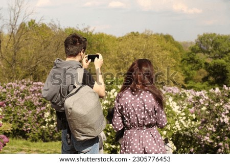 A guy and a girl take pictures on a mobile phone of the beauty of flowering lilac bushes on a blurred background. A loving couple is resting in nature. Memory of wonderful events
