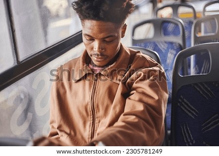 A young African man is riding a bus in downtown johannesburg wearing casual clothing and using his phone and drinking a ice coffee