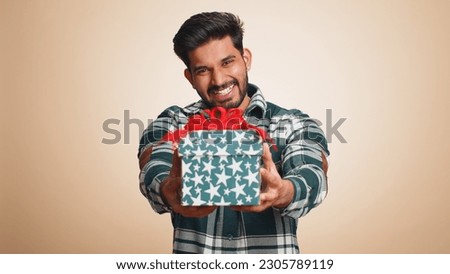 Positive smiling young indian man presenting birthday gift box stretches out hands, offer wrapped present career bonus, celebrating party, sale. Bearded hindu guy isolated on beige studio background