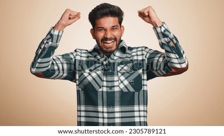 Happy joyful indian man shouting, raising fists in gesture I did it, celebrating success, winning, birthday, lottery jackpot goal achievemen, good news. Young hindu guy on beige studio wall background