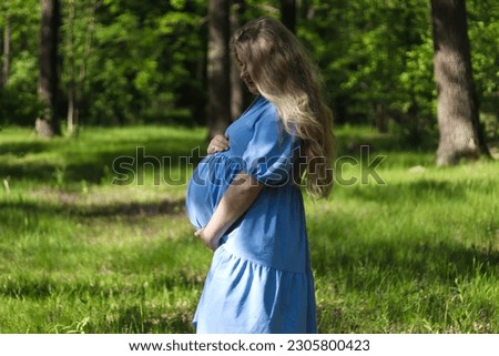 Beautiful young pregnant woman holding her belly against green forest lawn. Selective focus, outdoors. Concept of pregnancy and maternity