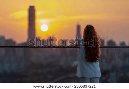 Woman looking and enjoying sunset view from balcony with the sun setting behind skyscraper in busy urban downtown with loneliness for solitude, loneliness and dreaming of freedom lifestyle Royalty-Free Stock Photo #2305837221