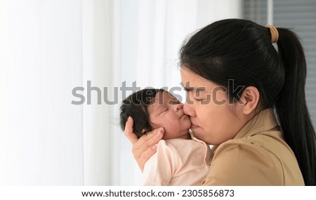 Asian mother holding newborn baby girl in her hands and gently kissing with love. Happy mom standing next to white curtain window at home. Safe with mom hug and healthy newborn baby concept