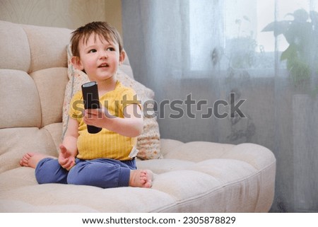 The little child sits on the sofa, watching TV with a remote control in hand and a smile on their face. The happy baby is watching their favorite show on TV, comfortably seated on the beige sofa.