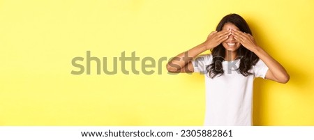 Image of excited african-american girl waiting for surprise, smiling and covering eyes with hands, standing over yellow background.