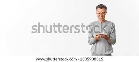 Image of happy, attractive middle-aged man in glasses and grey sweater, celebrating his birthday, smiling and looking at b-day cake, making a wish, standing over white background.