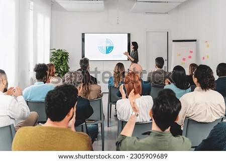 In a spacious office room, a businesswoman presents a SWOT analysis on a large screen. The diverse audience, viewed from the back, is clapping their hands in appreciation. Royalty-Free Stock Photo #2305909689