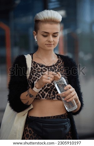 Diverse woman with short hair drinking water from a reusable glass bottle while listening to music