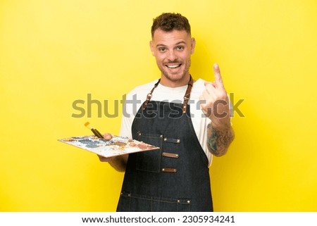 Young artist caucasian man holding a palette isolated on yellow background doing coming gesture