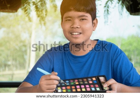 Young asian boy in blue t-shirt holding paintbrush and watercolor briquettes, sitting and drawing watercolor pictures in his freetime at home, teenager actitity and summer vacation concept.