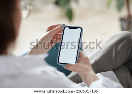 Mock up white screen blank mobile phone in woman hands holding sitting on armchair at home. Back view Royalty-Free Stock Photo #2306017185