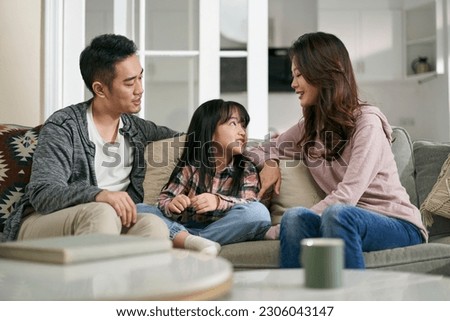 young asian mother and father sitting on family couch at home having a pleasant conversation with daughter