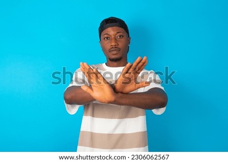 young man wearing striped T-shirt over blue studio background has rejection expression crossing arms and palms doing negative sign, angry face.