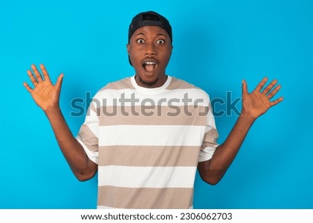 Optimistic young man wearing striped T-shirt over blue studio background raises palms from joy, happy to receive awesome present from someone, shouts loudly,