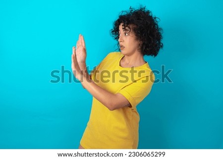 Displeased young arab woman wearing yellow T-shirt over blue background keeps hands towards empty space and asks not come closer sees something unpleasant
