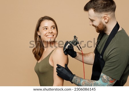 Side view happy tattooer master artist tattooed man wears green t-shirt apron hold machine black ink in jar equipment for making tattoo art on woman female body hand isolated on plain beige background