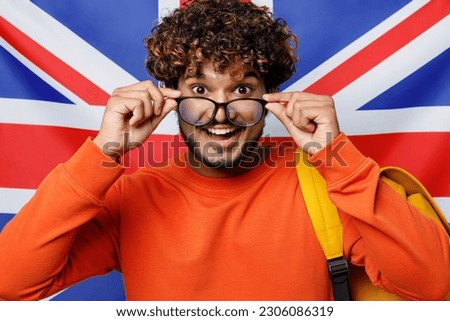 Young surprised shocked excited fun teen Indian boy student wearing casual clothes backpack bag lower glasses looking camera isolated on British flag background. High school university college concept