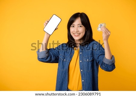 Happy asian young woman wearing yellow t-shirt denim shirt holding digital crypto currency coin and smartphone isolated on yellow background. Digital currency financial concept.
