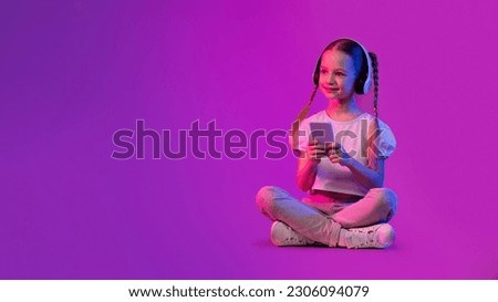 Positive cheerful cute preteen girl wearing summer outfit sitting on floor over luminous colorful background, using wireless headphones and smartphone, looking at copy space and smiling, web-banner