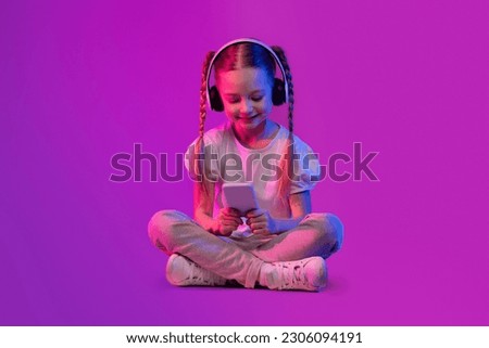 Digital Native, Generation Alpha Concept. Preteen kid happy pretty adorable girl in summer outfit sitting on floor over futuristic background, using wireless headphones and smartphone, copy space Royalty-Free Stock Photo #2306094191