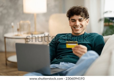 Ecommerce. Cheerful Middle Eastern Guy Enjoying Online Shopping Making Payment, Holding Credit Card And Using Laptop Computer Lying On Sofa At Home. Bank Offer Concept. Selective Focus