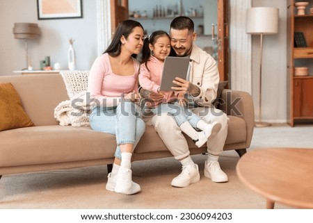 Family Online Leisure. Korean Parents And Little Daughter Using Tablet Watching Cartoons Online On Computer Sitting Together In Modern Living Room Indoor. Internet And Fun Concept