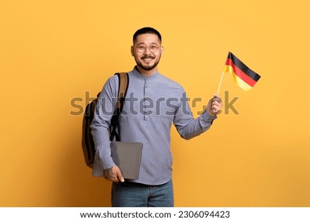 Online Language Courses. Smiling Young Asian Man Holding German Flag And Laptop While Standing Over Yellow Background In Studio, Millennial Man Recommending Remote Education Programs, Copy Space Royalty-Free Stock Photo #2306094423