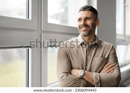 Positive middle aged businessman standing near window in company office, looking away and smiling, copy space. Successful man entrepreneur posing with crossed arms Royalty-Free Stock Photo #2306094443