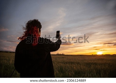 woman red hair taking mobile phone pictures of the sunset with purple, purple, orange, and blue colors, at the sunset