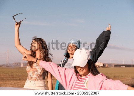 The three European friends traveling in a white convertible car during the summer vacations have paused on the road and raise their arms cheerfully.