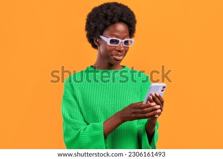 African American young woman with afro hair styling standing in a green sweater on a bright orange background wearing glasses and using the phone.