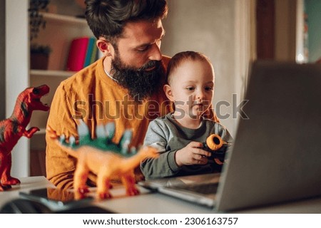 A happy family is spending time together. A freelancer is taking a break from work and spending time with his adorable son at the home office. A freelancer and his toddler watch cartoons together.