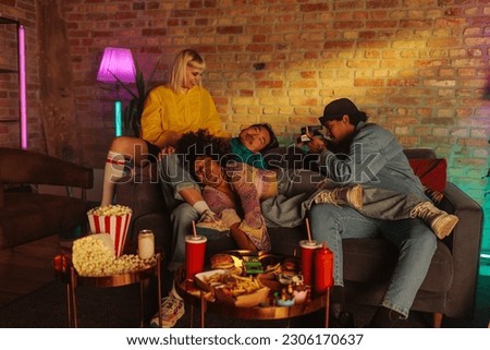 A group of young stylish people are at home, sleeping after eating a big takeout meal and one of them is taking a polaroid picture of them.