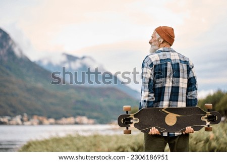 Active cool happy bearded old hipster man standing in nature park holding skateboard. Mature traveler skater enjoying freedom spirit and extreme sports hobby looking at mountains view.