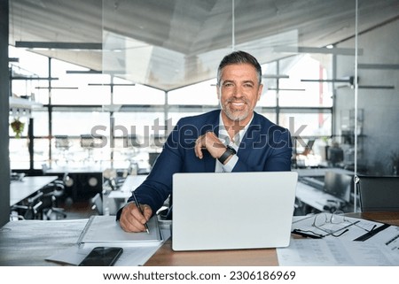 Happy middle aged professional business man company executive ceo manager wearing suit sitting at desk in modern office working on laptop computer and writing notes, portrait at workplace. Royalty-Free Stock Photo #2306186969