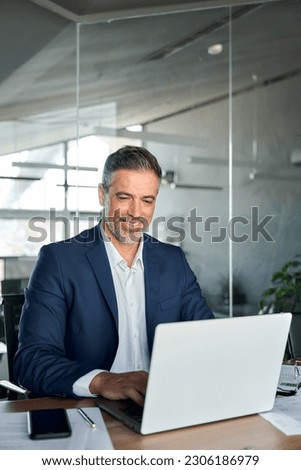 Happy middle aged business man ceo wearing blue suit sitting at desk in office using laptop. Mature businessman professional executive manager working on computer technology at workplace. Vertical Royalty-Free Stock Photo #2306186979